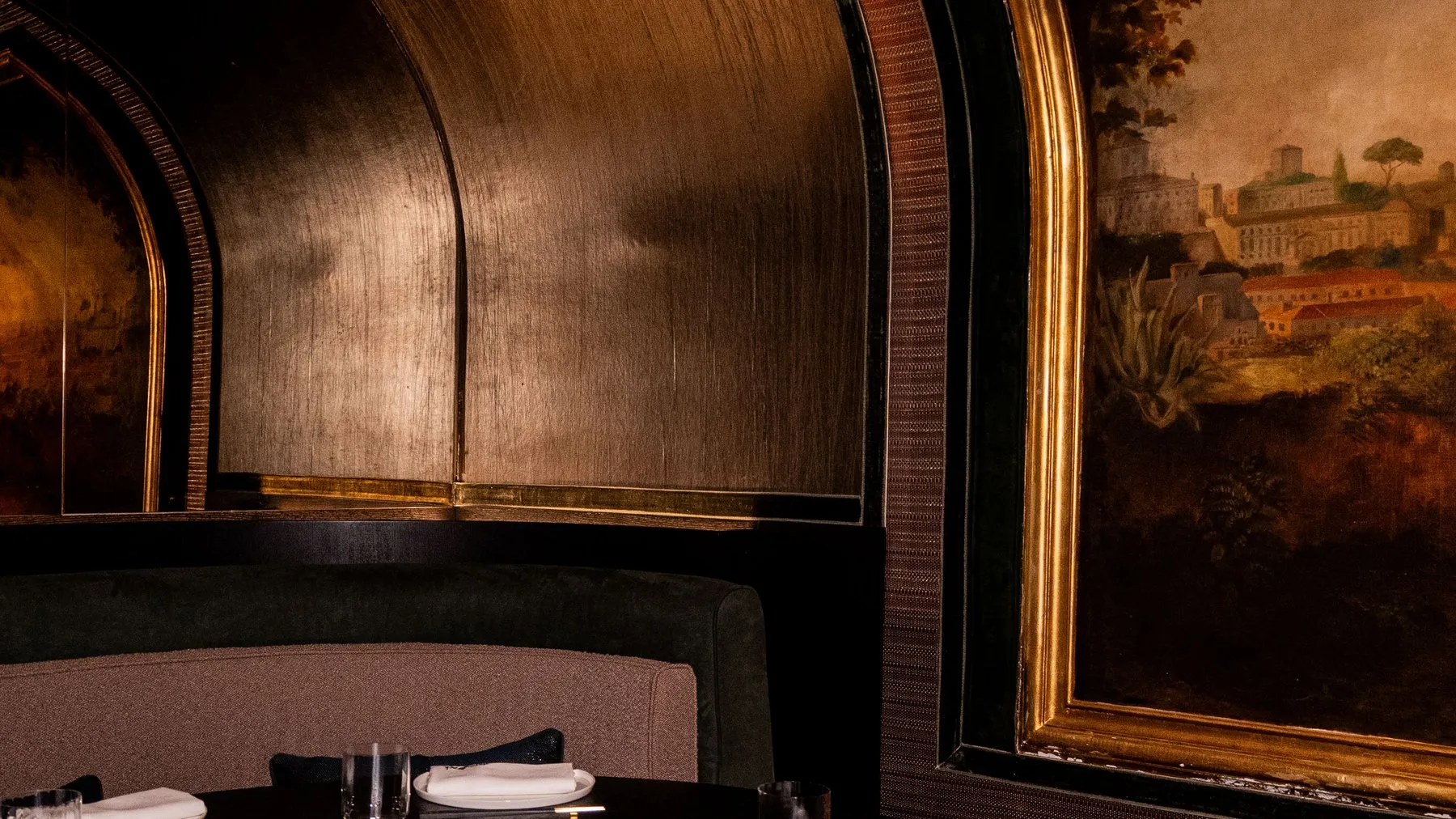 A round black table with a curved banquette under a gold vaulted ceiling, beside a gilded painting of a landscape