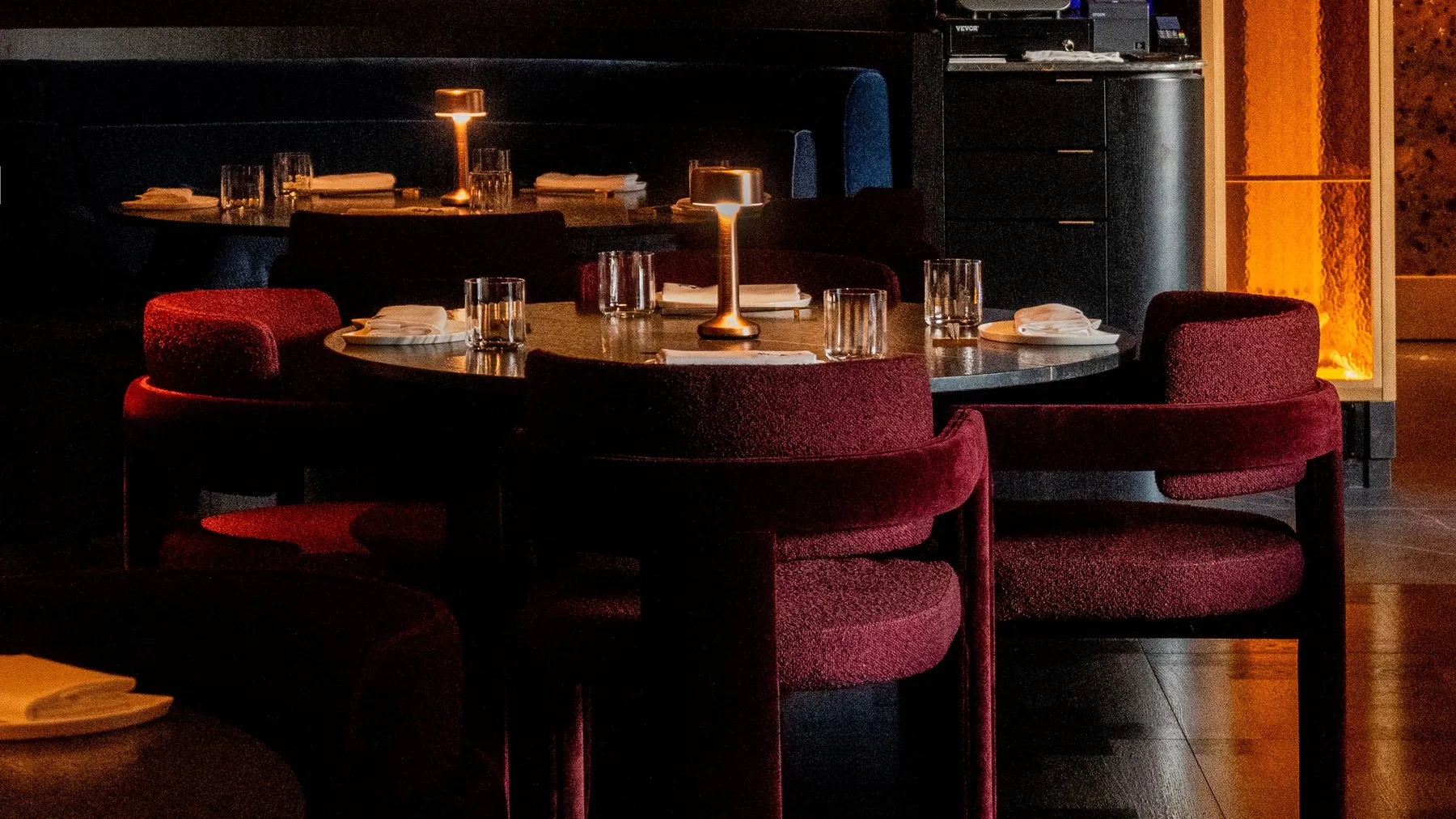 Round tables with deep red bouclé chairs and bronze table lamps against a copper wall