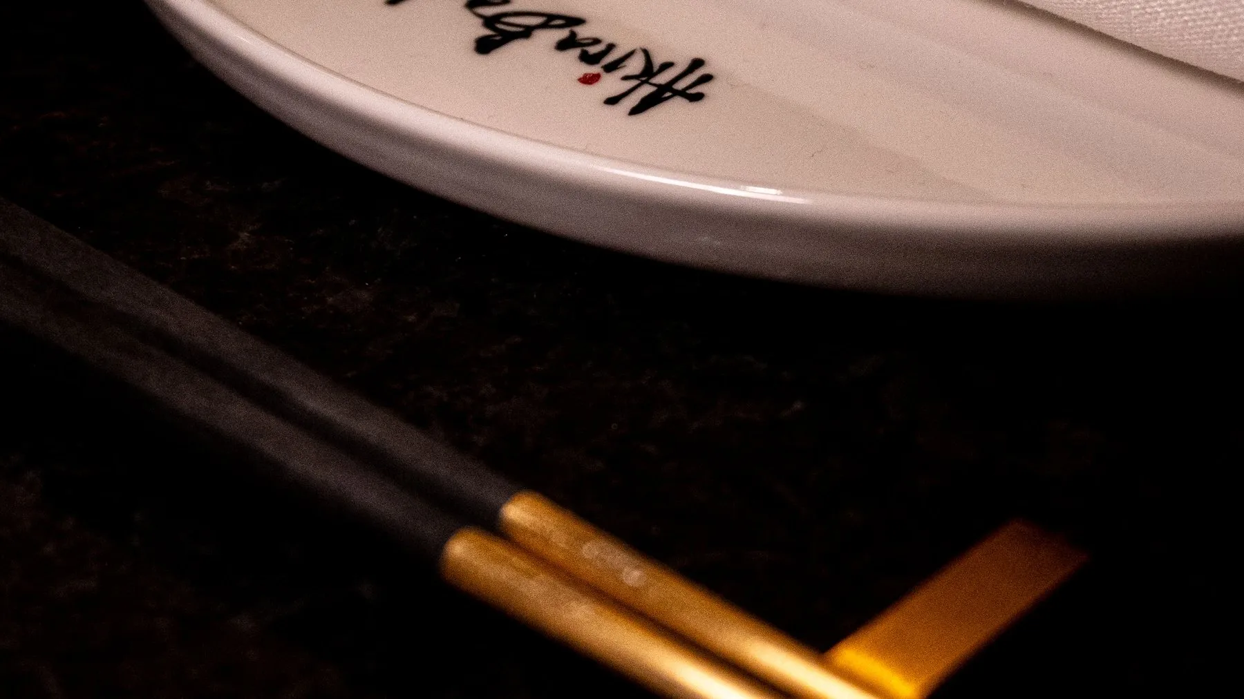 Gold-tipped chopsticks resting on a brass stand beside a plate printed with the Akira Back signature