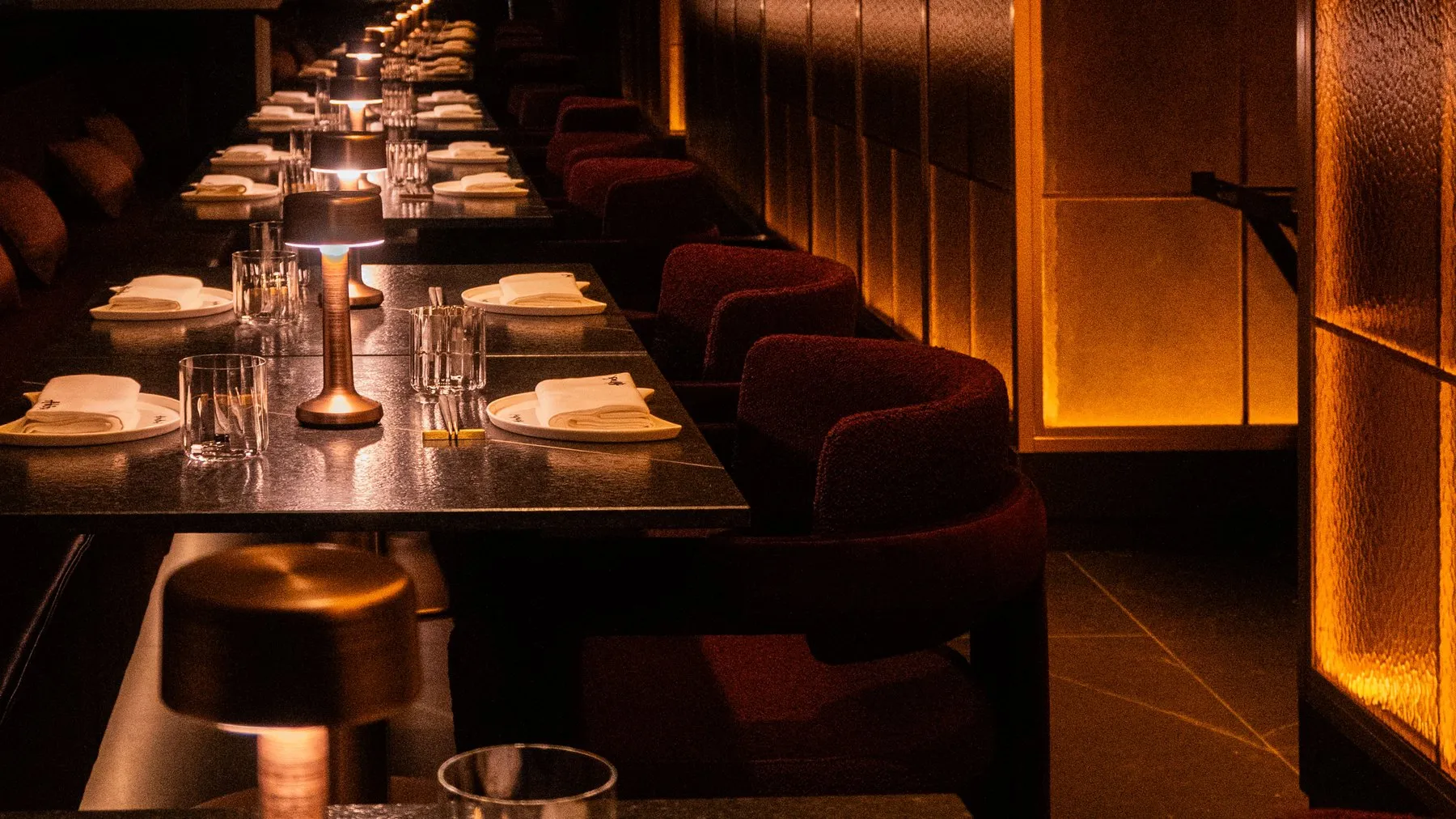 A long row of tables set with white napkins and bronze lamps, beside a wall of lit amber glass