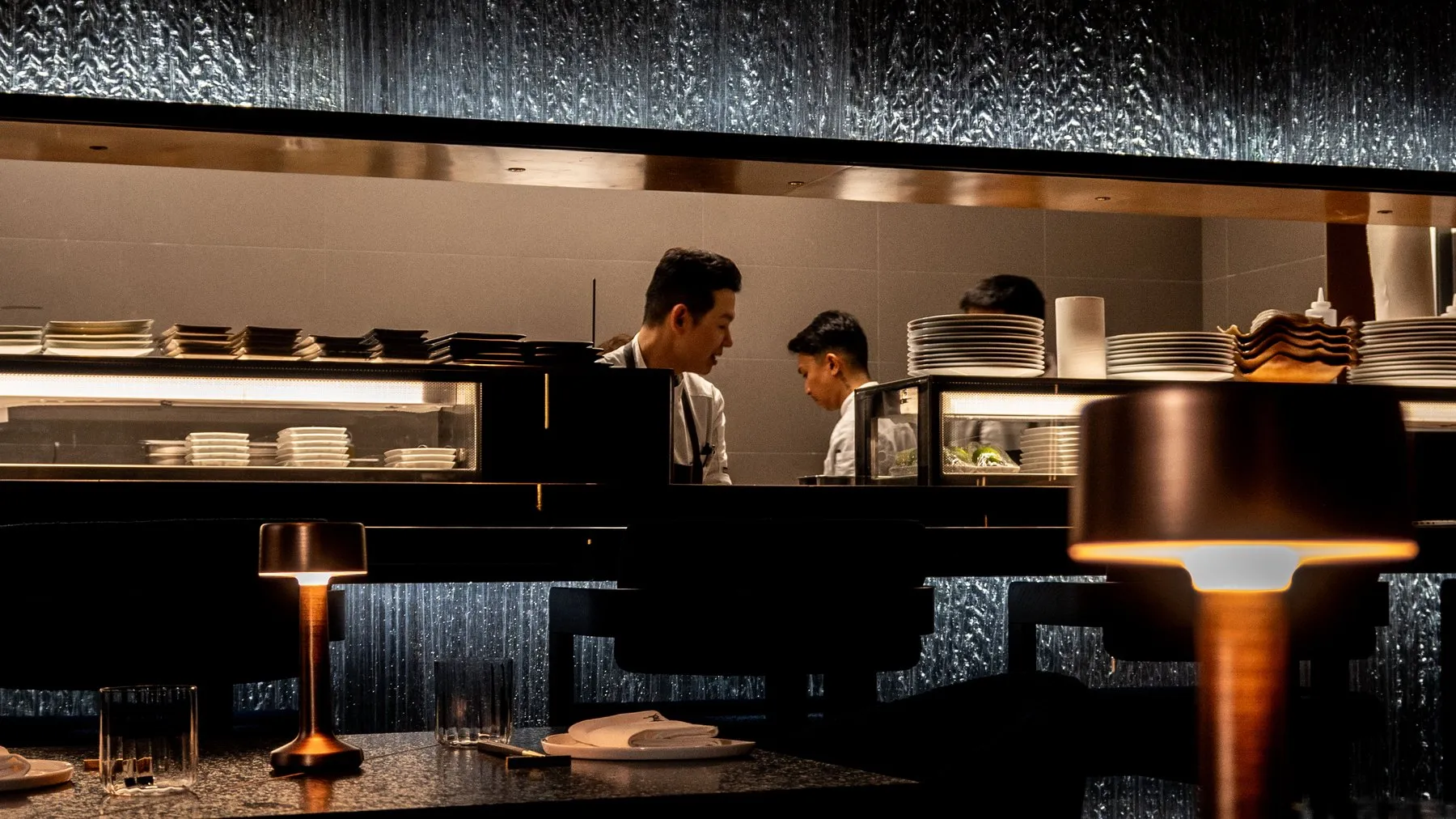 The open kitchen seen across the dining room, chefs at work under a wall of backlit rippled glass