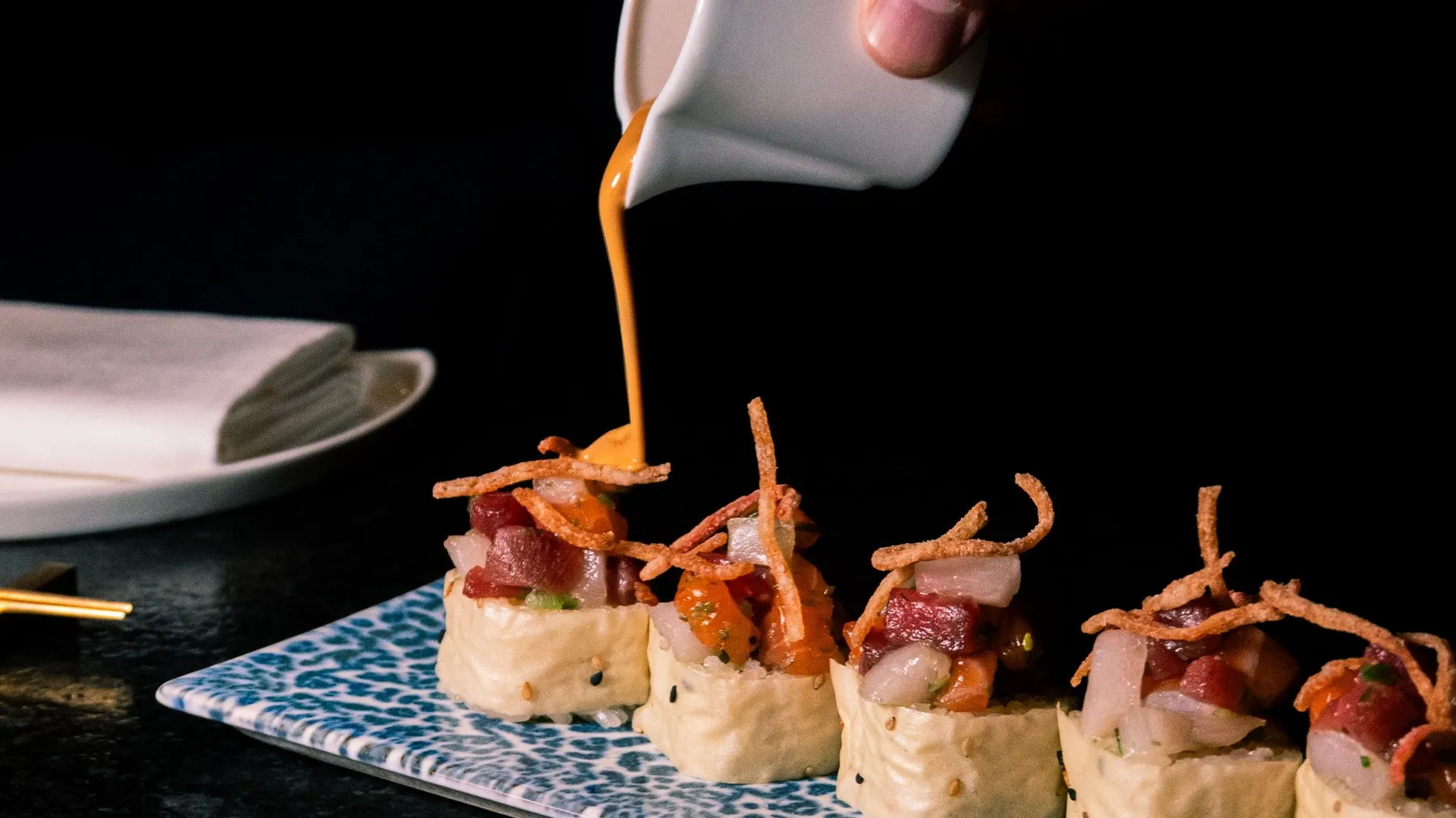 A hand pouring an orange sauce over a row of sushi rolls topped with diced tuna, salmon and crispy shallots