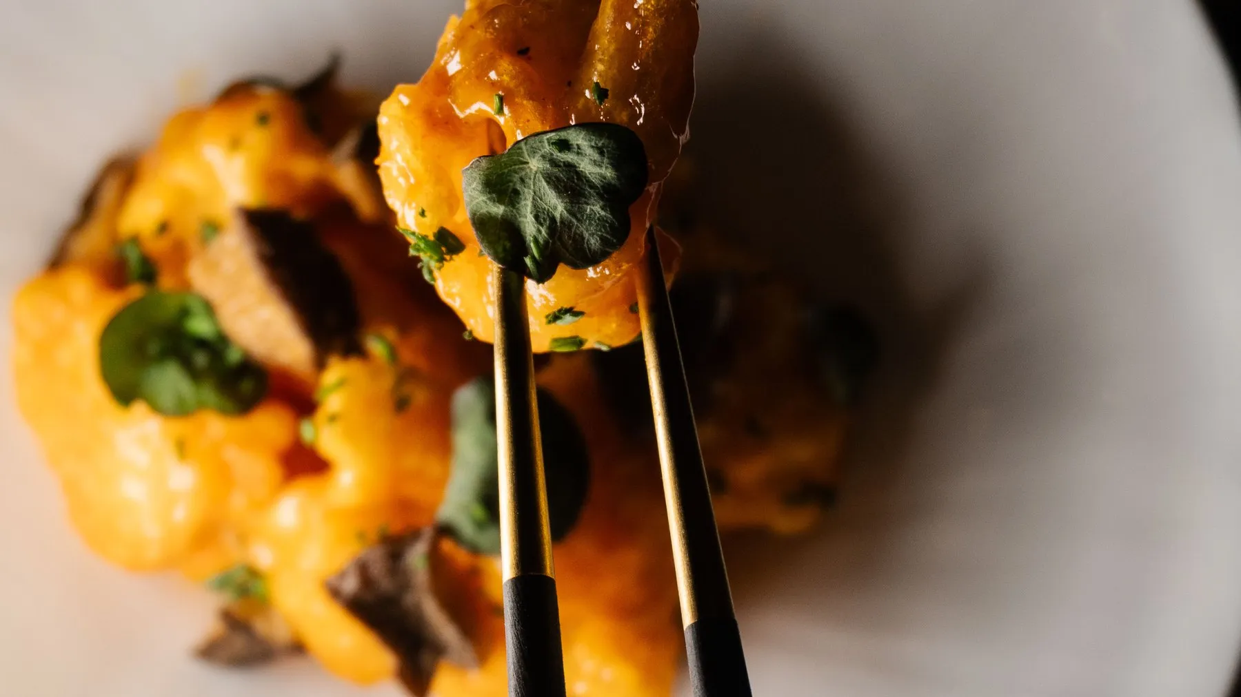Chopsticks lifting a glazed, sauce-coated bite from a white plate scattered with mushrooms and herbs