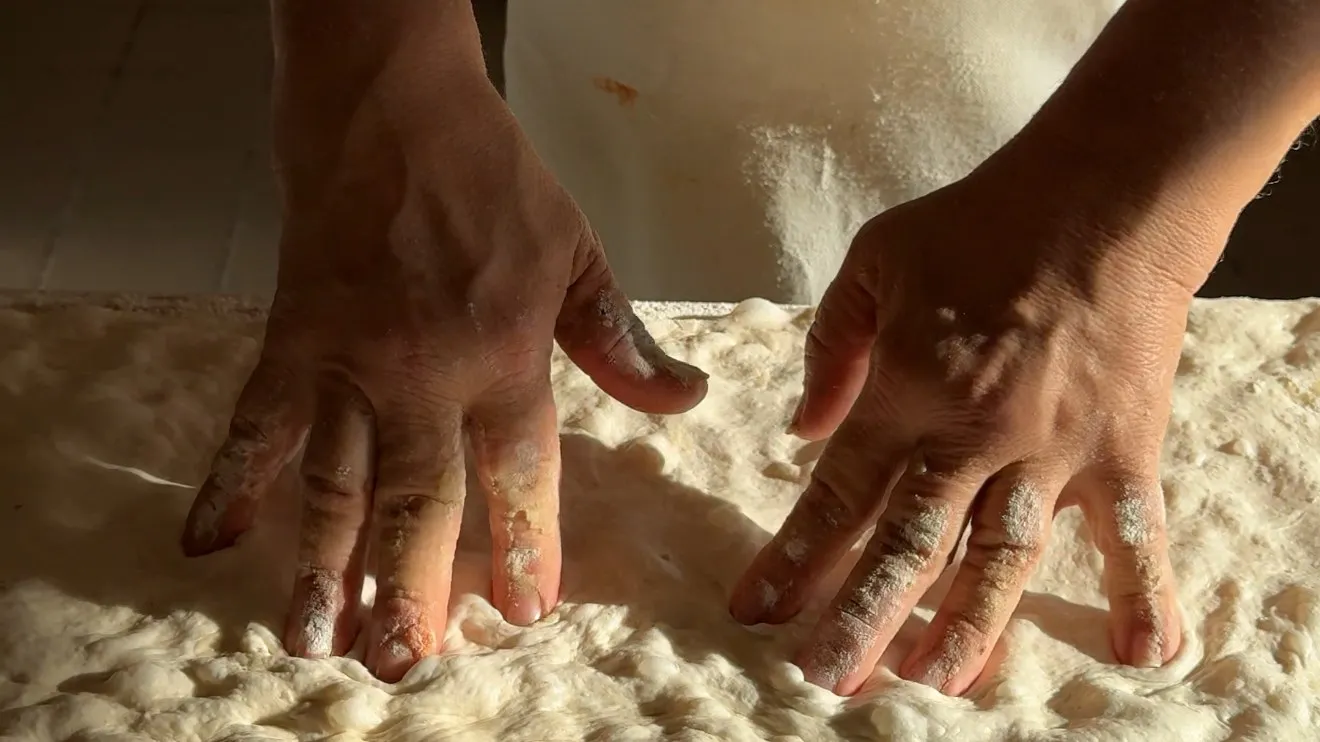 Two floured hands pressing dimples into a long sheet of pizza bianca dough on a wooden board