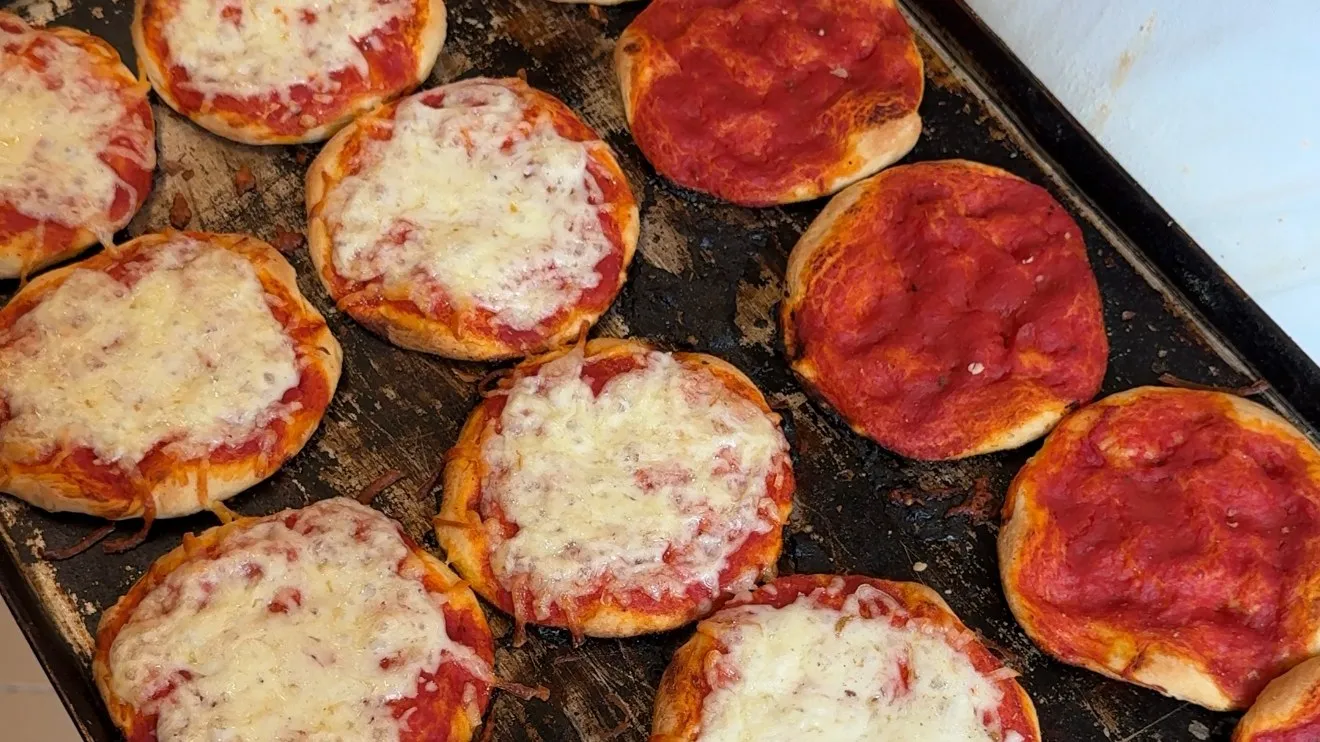 A baking tray of small round pizzette, most with tomato and melted cheese, a few with tomato only