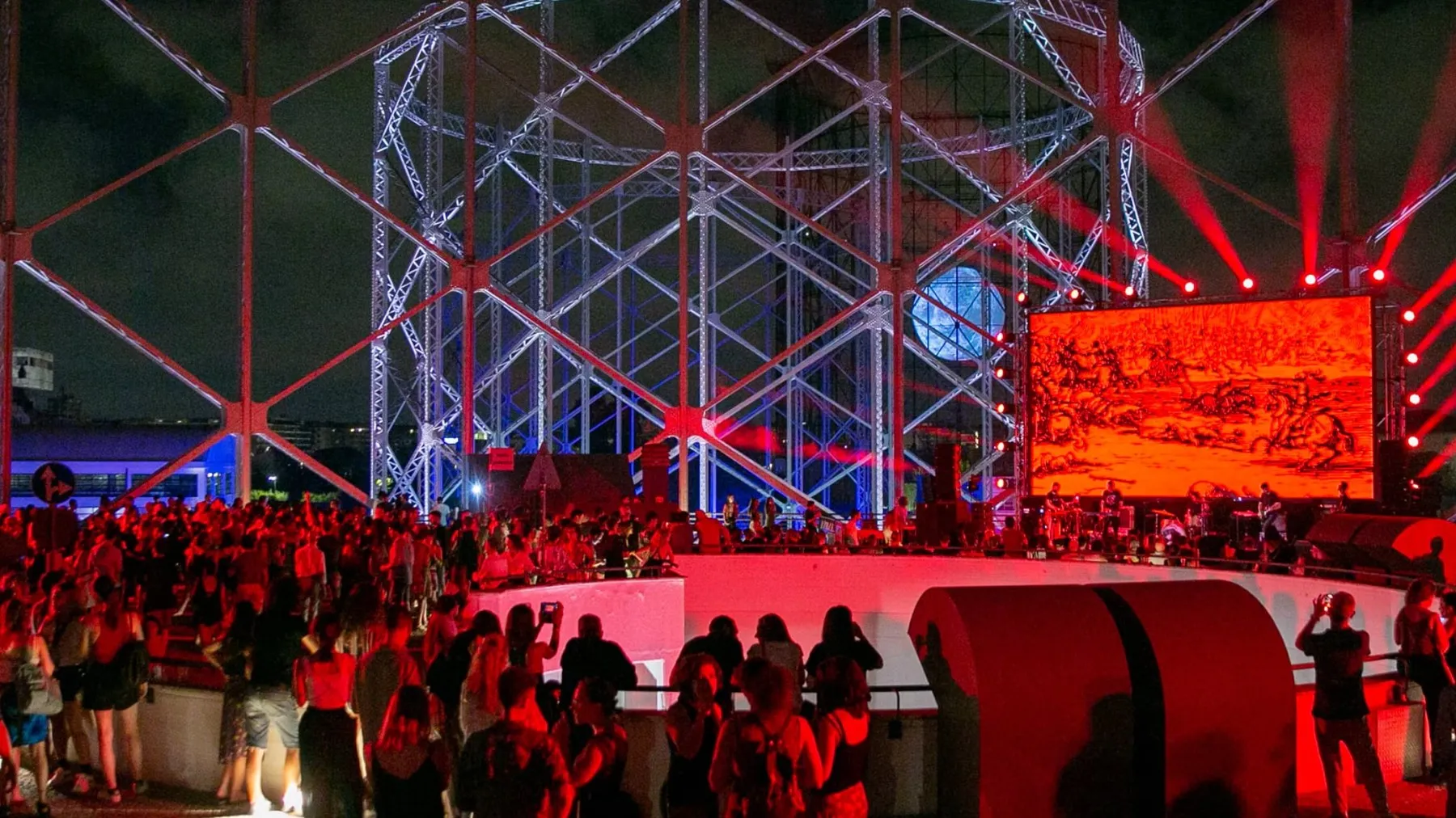 A crowd at night under the iron frame of the Gazometro, lit red, with a stage and a big screen