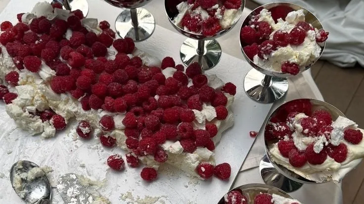 Silver coupes of meringue, cream and raspberries around a broken pavlova on a white tablecloth