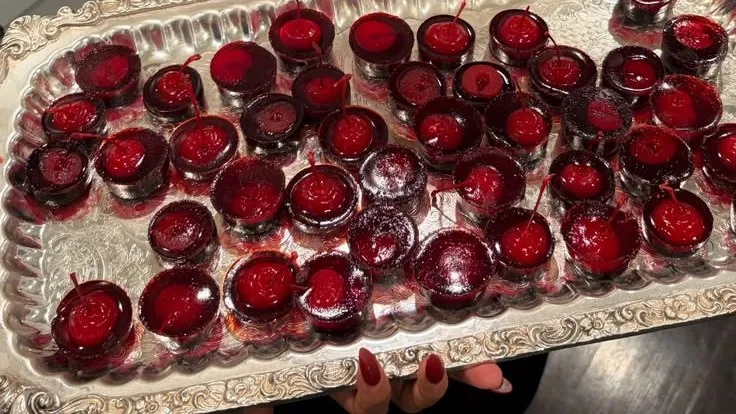A waiter in a bow tie holding a silver tray of blood-red jelly shots topped with cherries