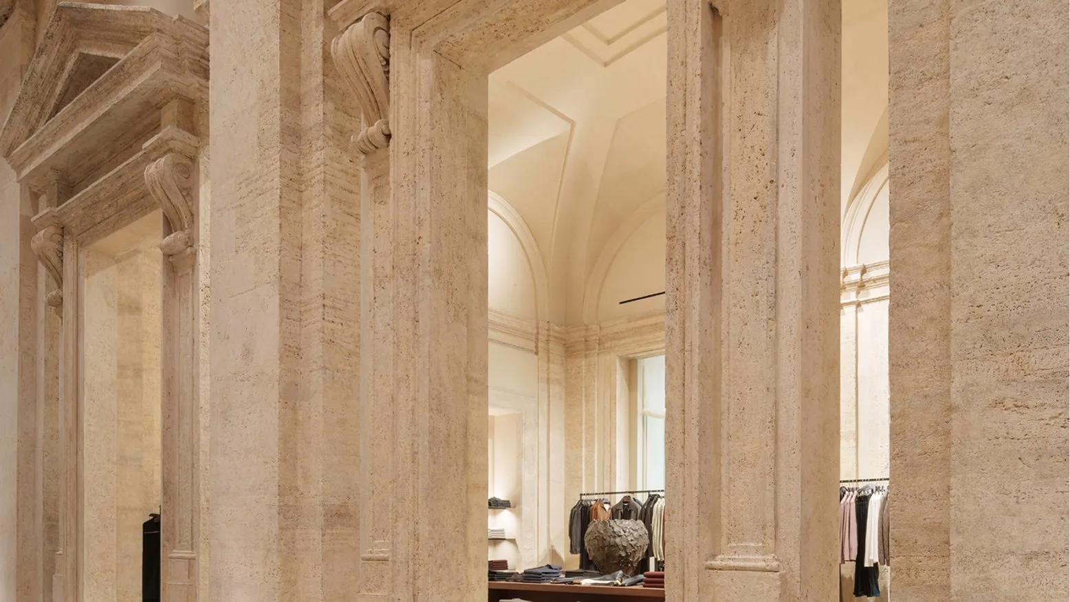 Travertine doorways with carved scrolls opening onto a room of clothing rails and a herringbone wooden floor
