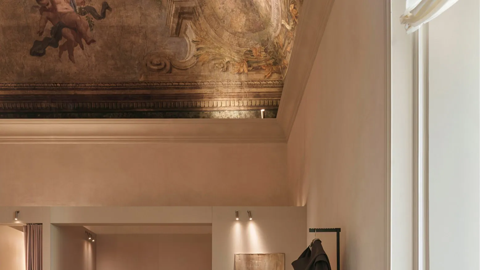A fitting area with a pink linen bench and a coat on a stand, under a painted ceiling with cherubs and garlands