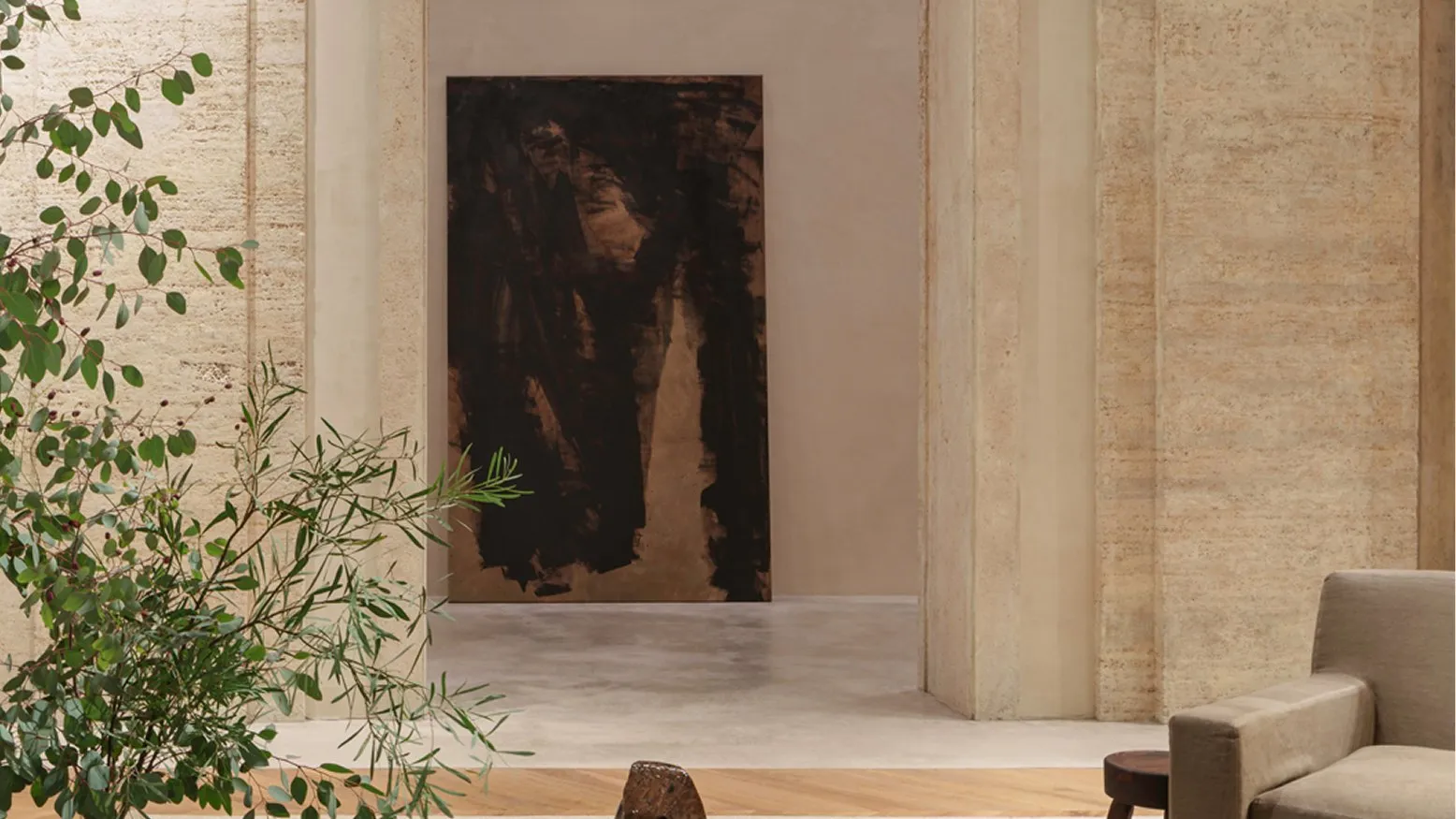 A sitting area with a long slatted wooden table and a linen armchair, in front of a travertine doorway with a dark painting beyond