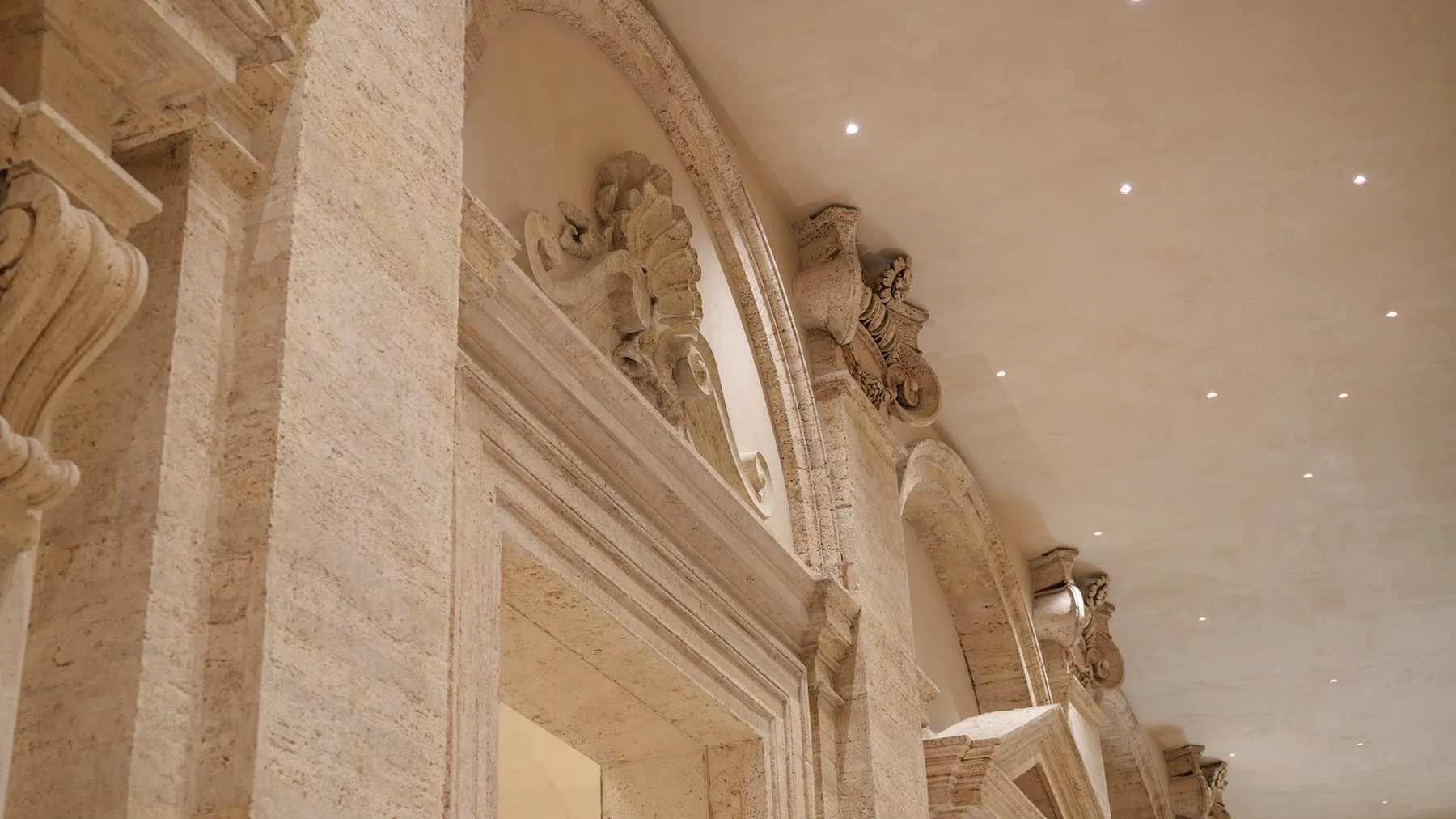Carved stone pediments and arched lunettes along a travertine wall, under a ceiling set with small spotlights