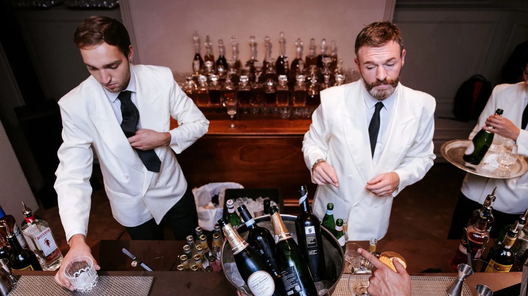 Two bartenders in white jackets behind a bar with an ice bucket of champagne bottles