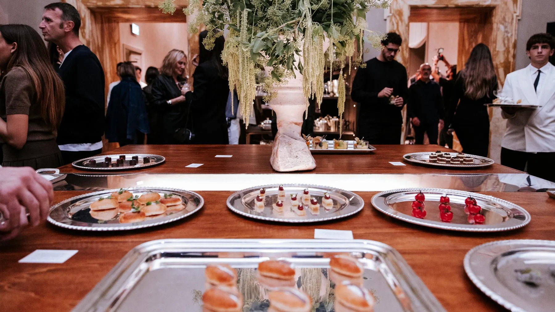A wooden table of silver trays with small bites and a tall flower arrangement, guests mingling behind