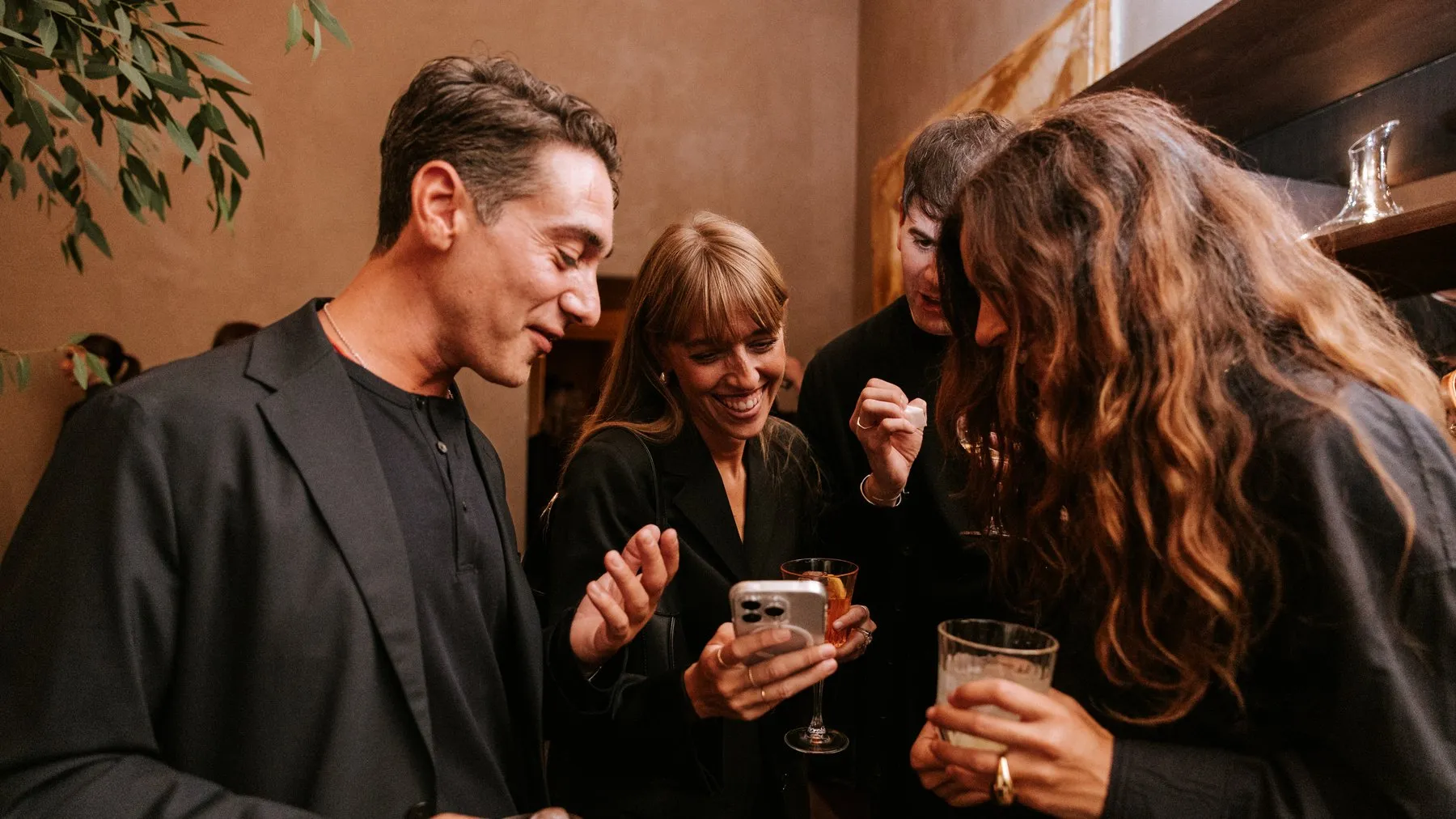 A group of guests laughing over a phone, drinks in hand