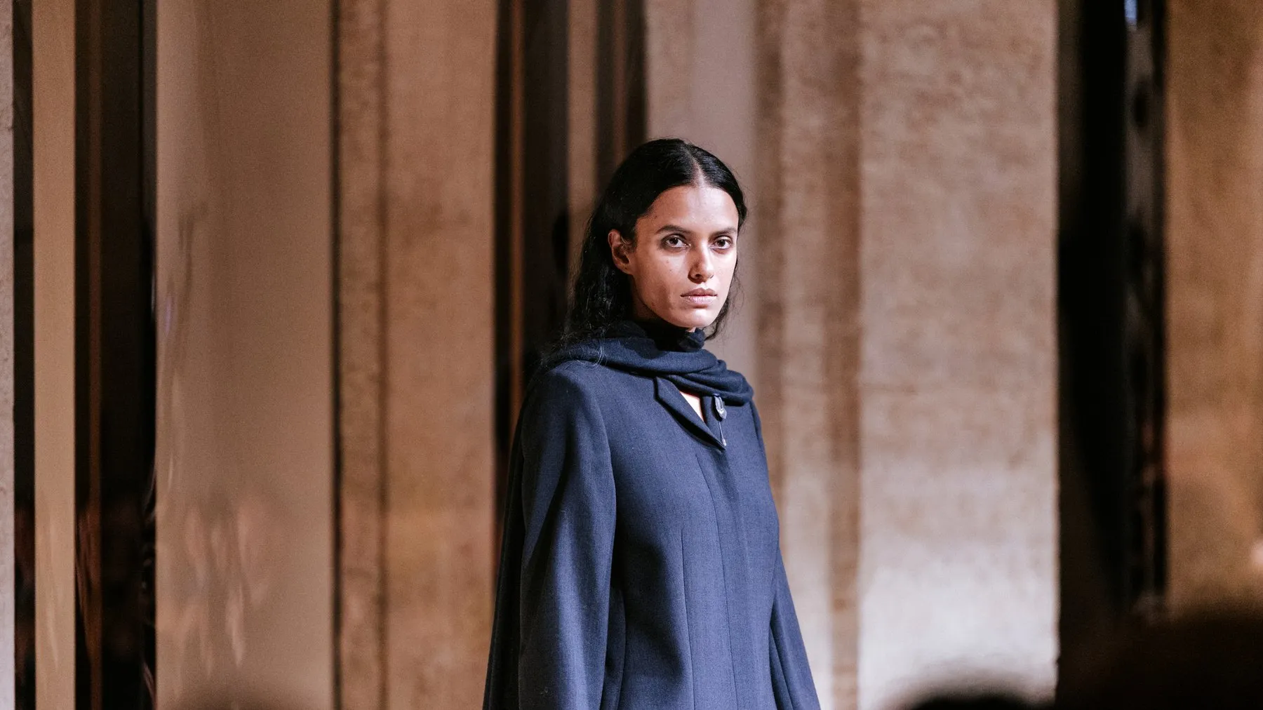 A model in a long tailored navy coat with a scarf wrapped high at the neck