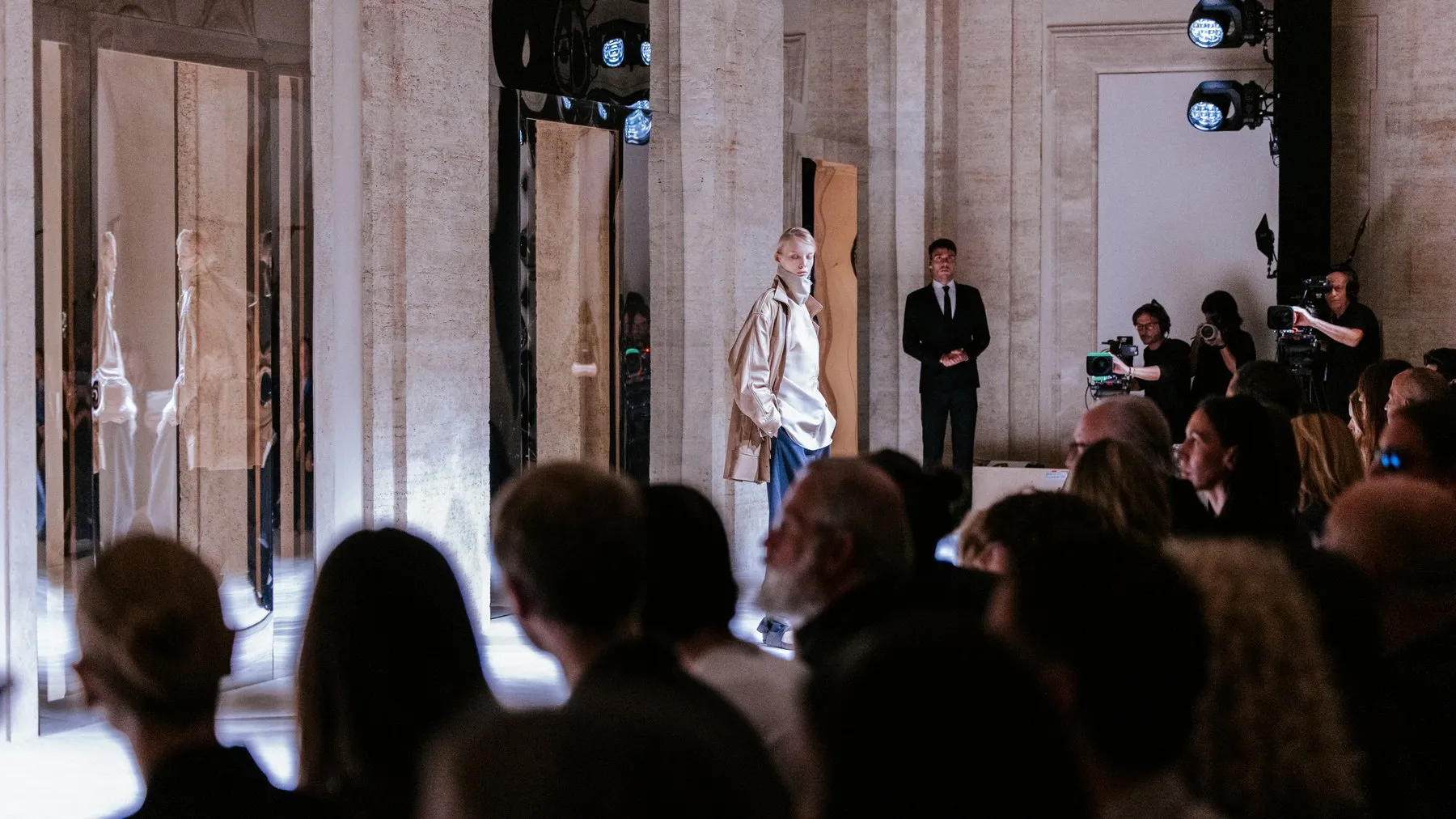 A model in a pale leather jacket over a white shirt and wide blue trousers, reflected in a mirrored panel