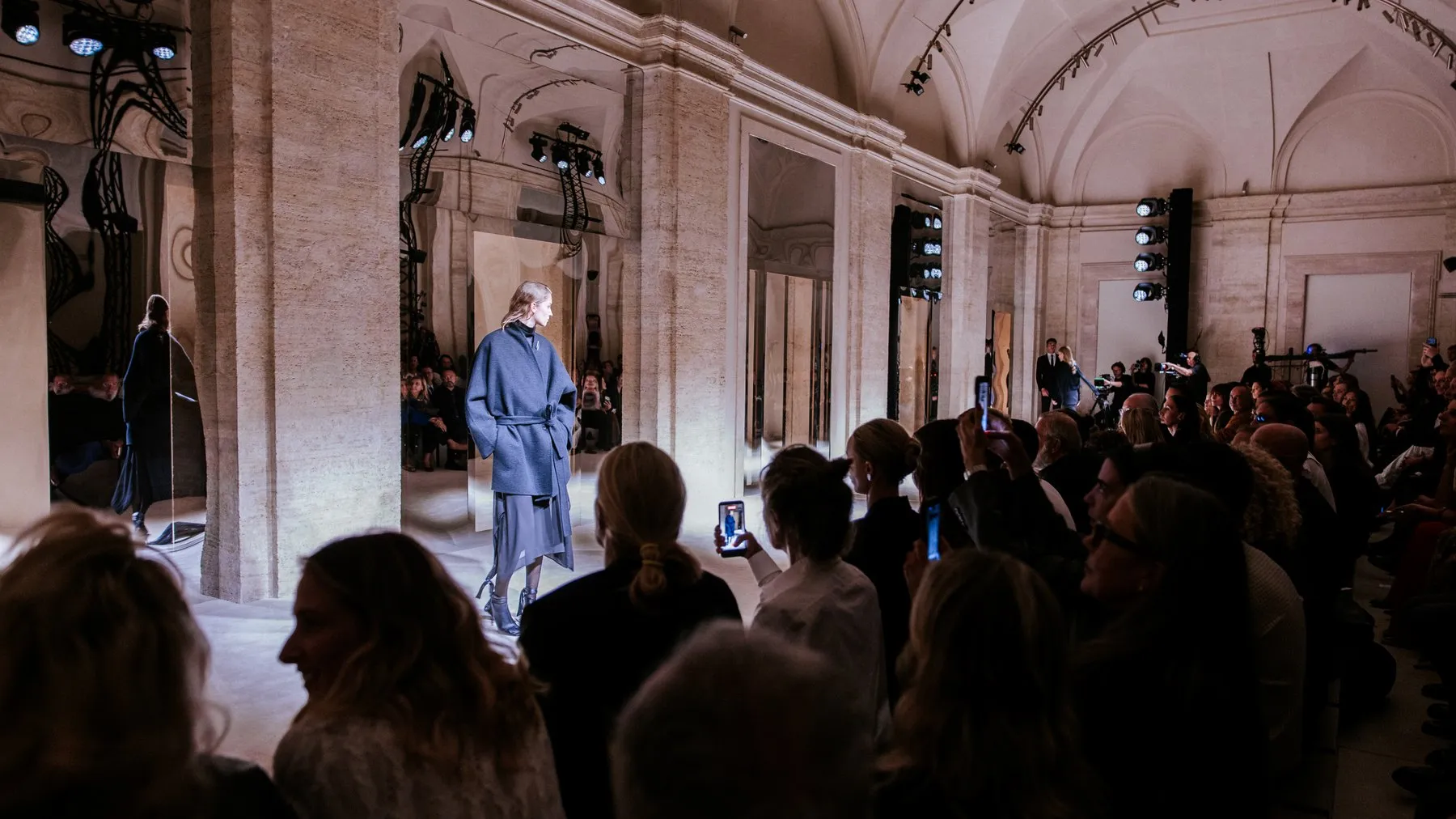 A model in a belted grey-blue coat walking between travertine pillars and mirrored panels, watched by a seated crowd